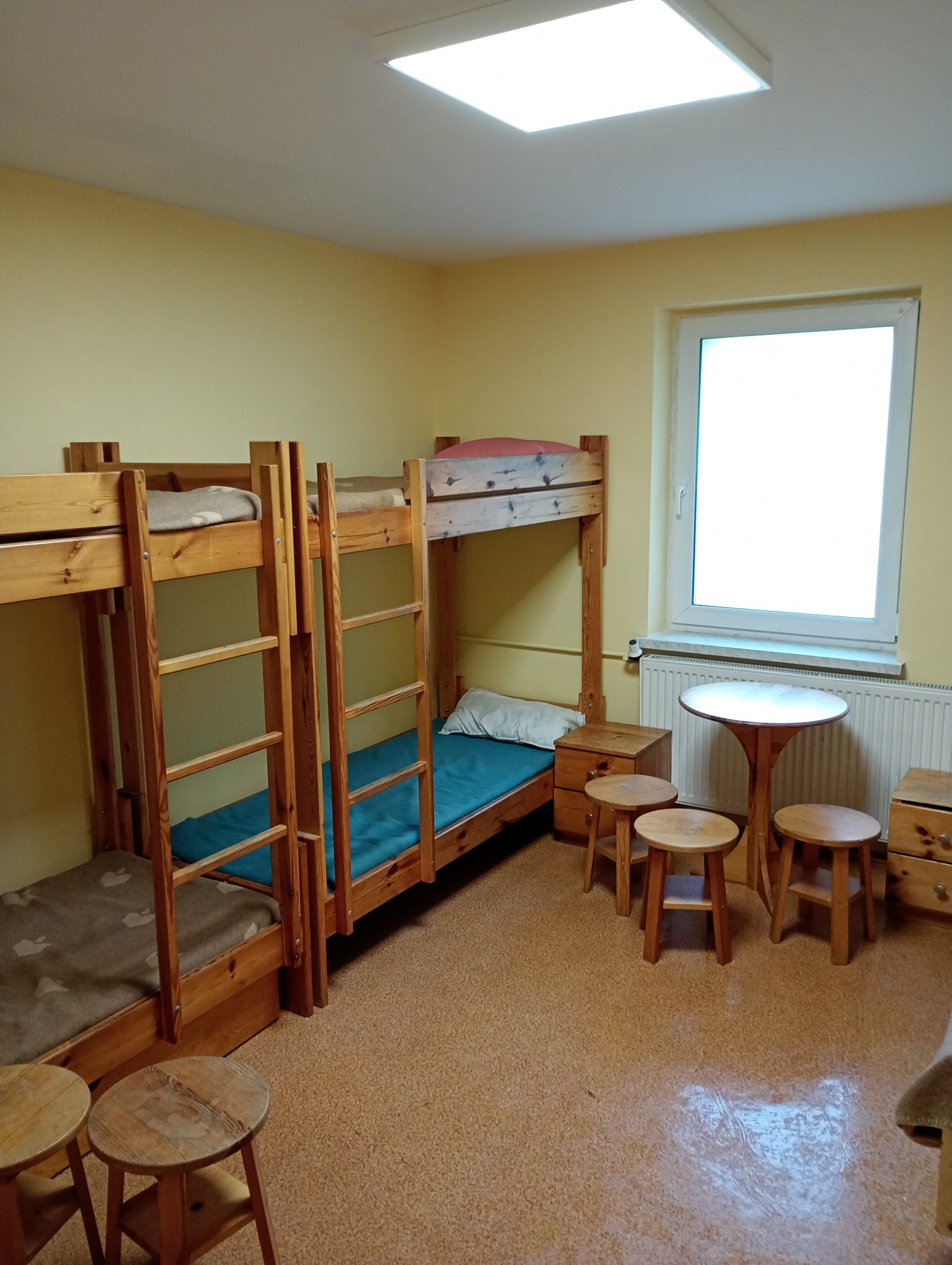Prosto urządzony pokój z dwoma drewnianymi łóżkami piętrowymi, małymi stolikami nocnymi i okrągłym stolikiem z trzema taboretami, oświetlony przez lampę sufitową i światło dzienne z okna.