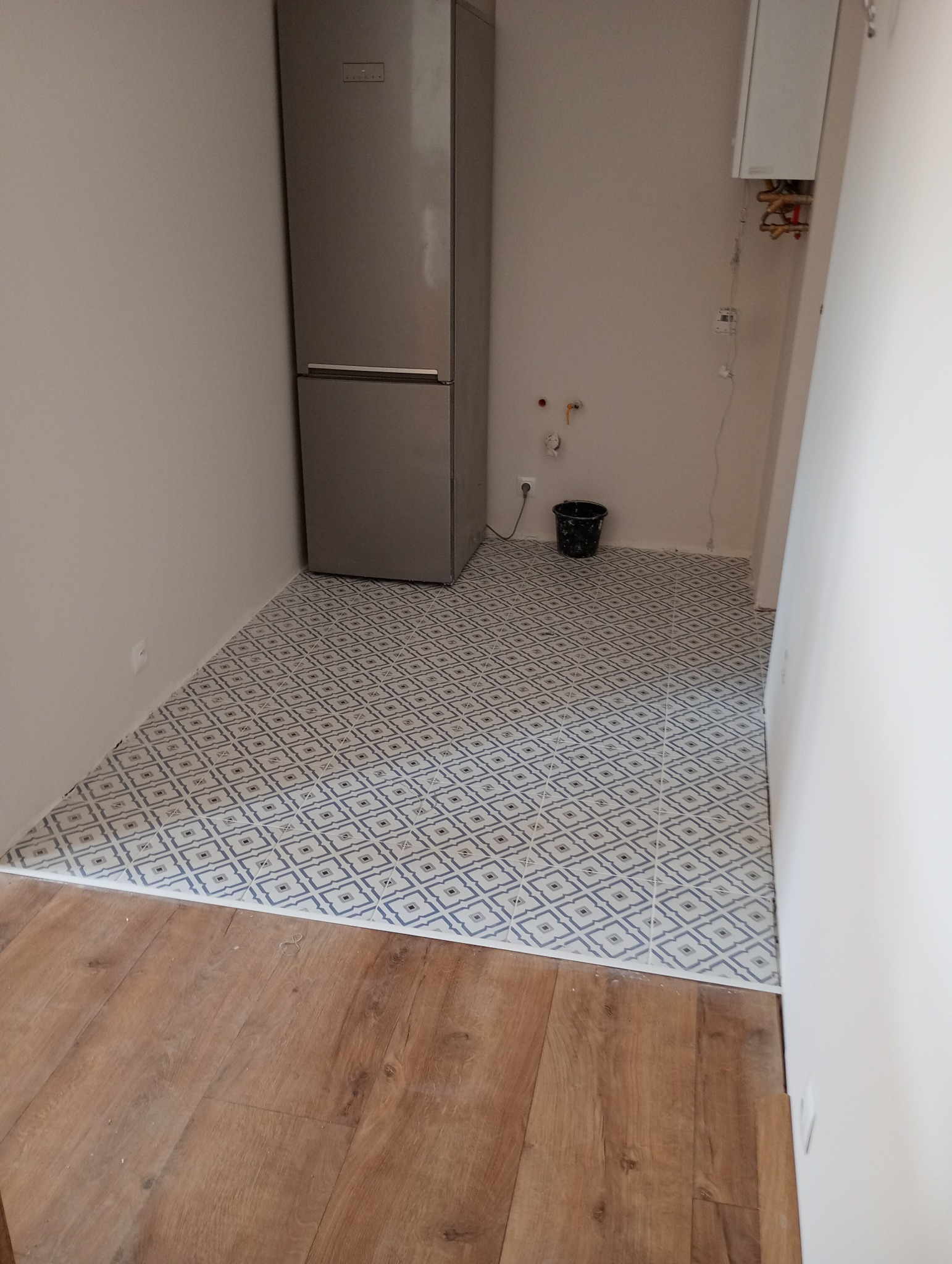 Nowo wyłożona podłoga w kuchni z płytek w stylu marokańskim, kontrastująca z drewnianą podłogą w sąsiednim pomieszczeniu, widoczna lodówka i instalacja wodna na ścianie.