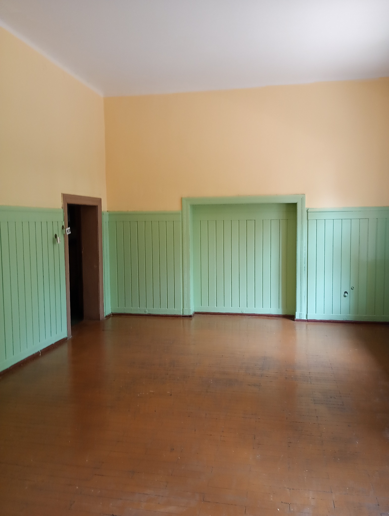 Puste pomieszczenie przed remontem: parkiet z widocznymi śladami użytkowania, ściany pomalowane w górnej części na żółto, a w dolnej wykończone zieloną boazerią z pionowych paneli.