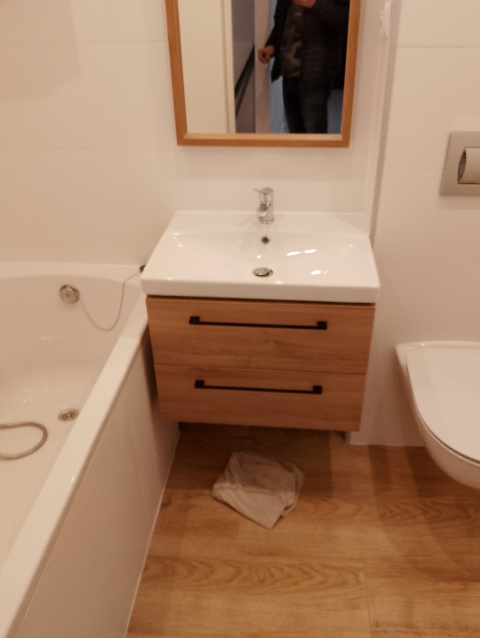 Nowoczesna łazienka z drewnianą szafką pod umywalką, białymi płytkami i wanną, widoczne odbicie osoby w lustrze.