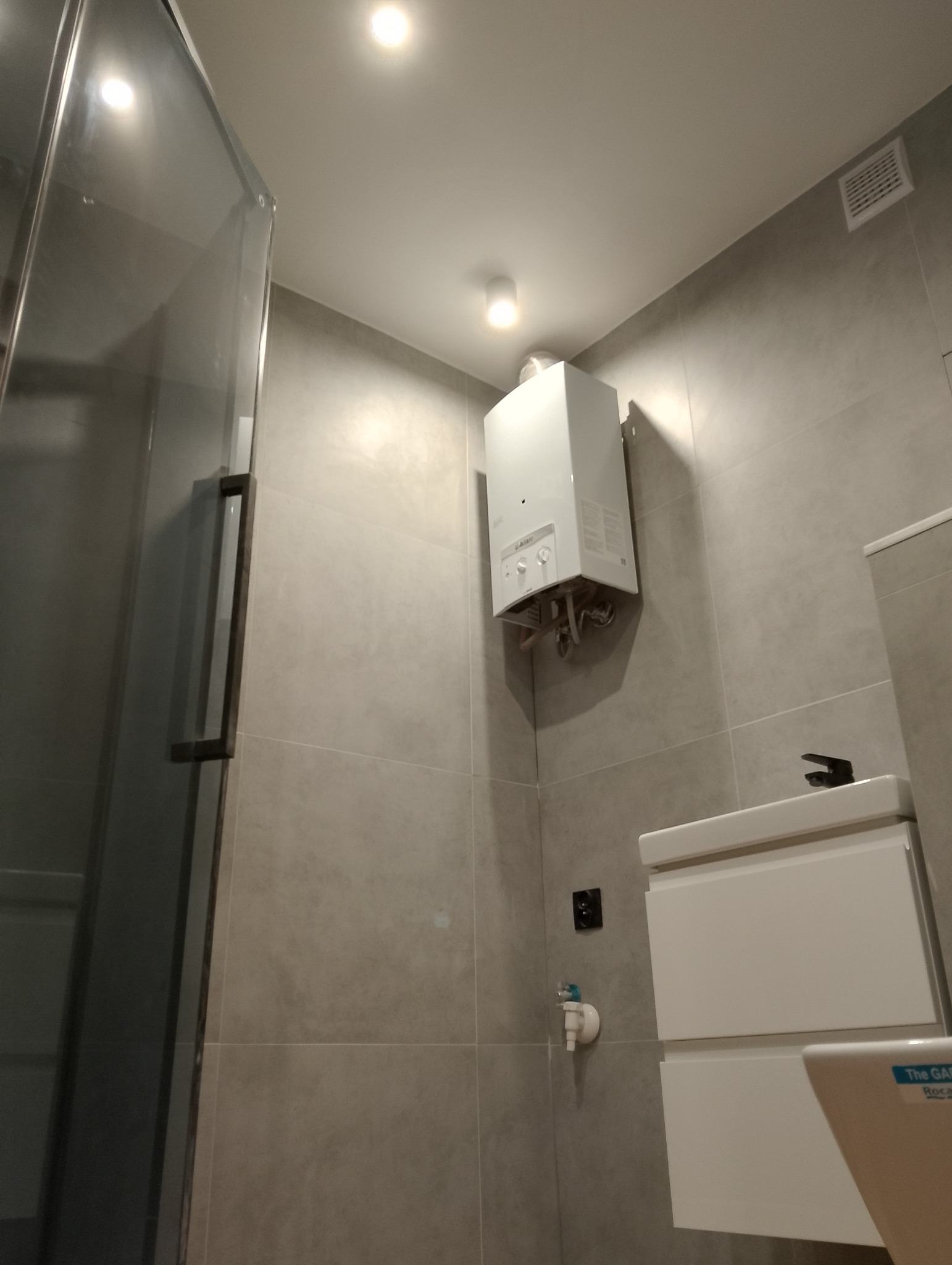 Wnętrze łazienki po remoncie: szara glazura, biała szafka z umywalką, wiszący piec gazowy, kabina prysznicowa z szklanymi drzwiami.