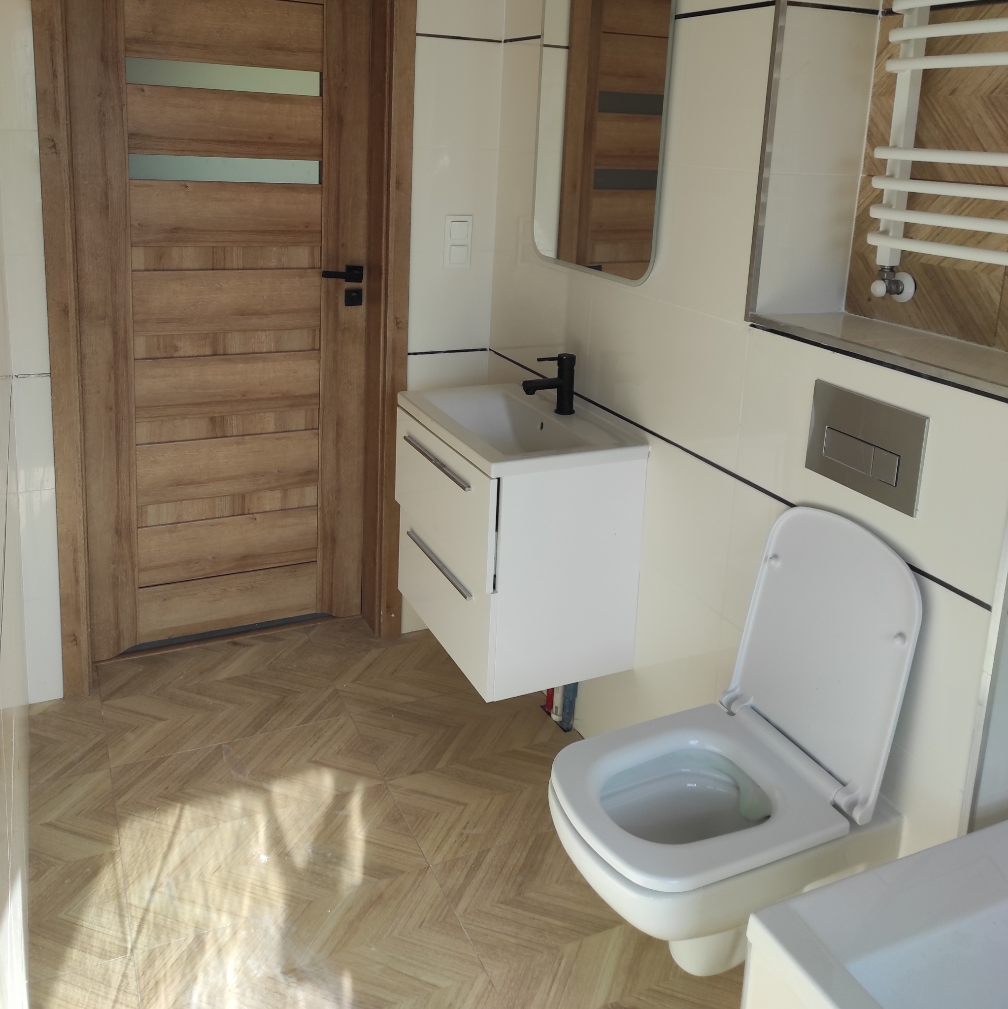 Nowoczesna łazienka z drewnianymi drzwiami, białą umywalką z szafką, toaletą i grzejnikiem drabinkowym na tle beżowych płytek z ciemnymi fugami.