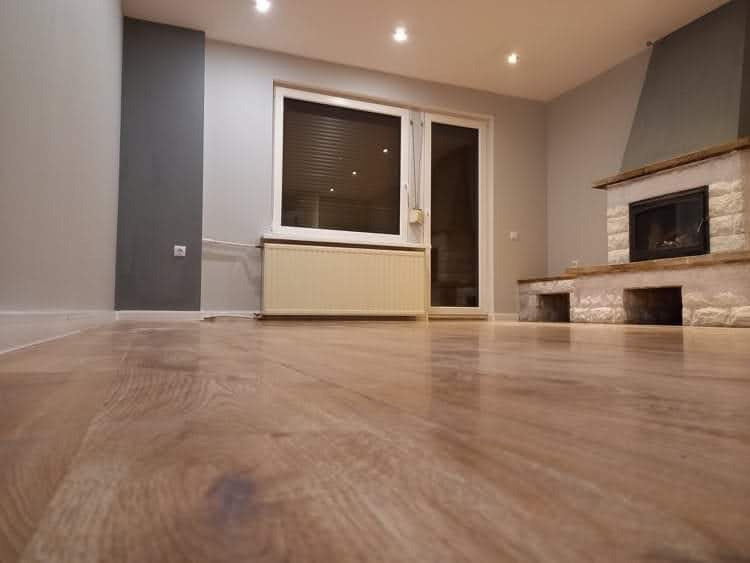 Jasne panele podłogowe w przestronnym salonie z kominkiem i grzejnikiem pod oknem. Ściany w odcieniach szarości, widoczne okno balkonowe i nowoczesne oświetlenie.