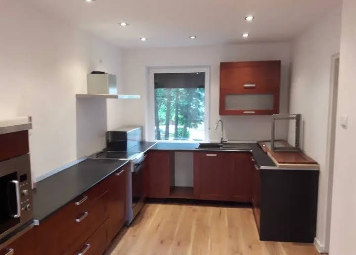 Nowoczesna kuchnia z drewnianymi frontami, czarnym blatem i parkietem. Okno z widokiem na zieleń. Górna szafka z przeszklonym frontem. Oświetlenie punktowe.