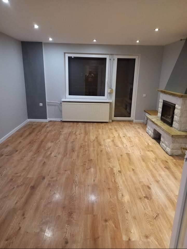 Jasne panele podłogowe w przestronnym pokoju z szarymi ścianami, białym oknem i kamiennym kominkiem. Widoczne detale wykończenia wnętrza.
