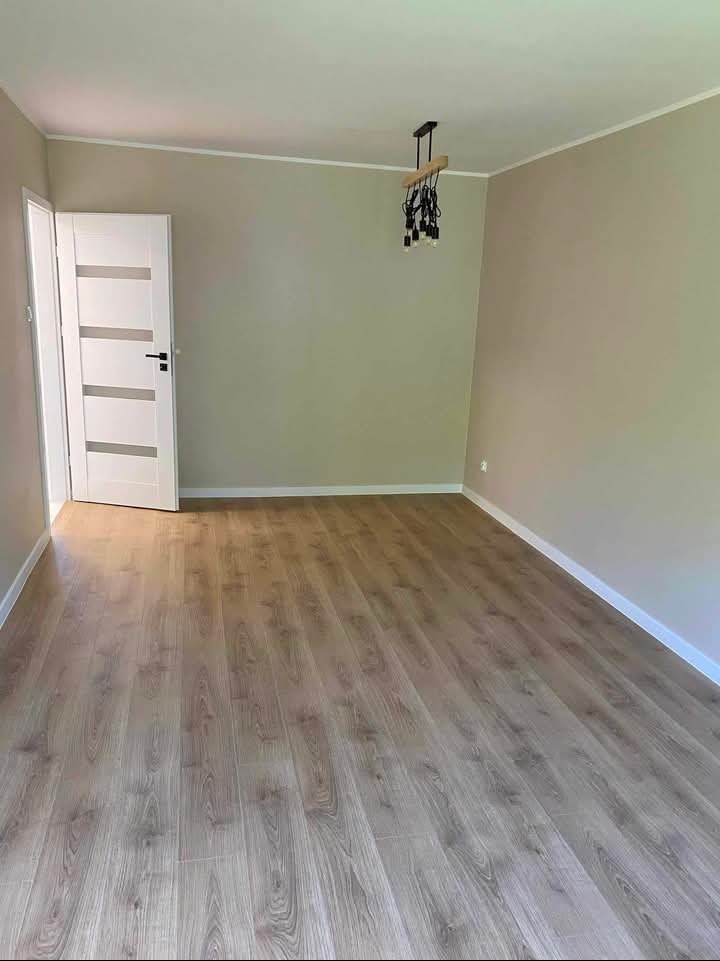 Jasne panele podłogowe w pokoju z beżowymi ścianami, białe listwy przypodłogowe i drzwi z przeszkleniami. Nowoczesna lampa wisząca.