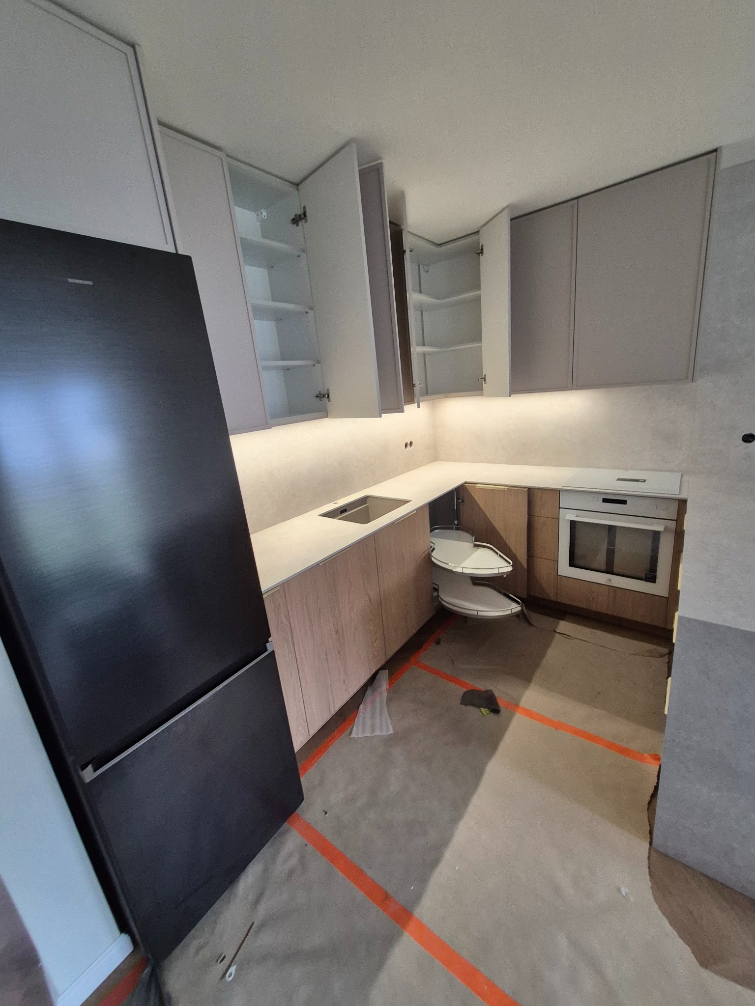 Nowoczesna kuchnia w trakcie montażu, widoczna zabudowa z szarymi i drewnopodobnymi frontami, otwarte szafki wiszące, wbudowany piekarnik, wysuwana półka narożna, czarna lodówka, podłoga...