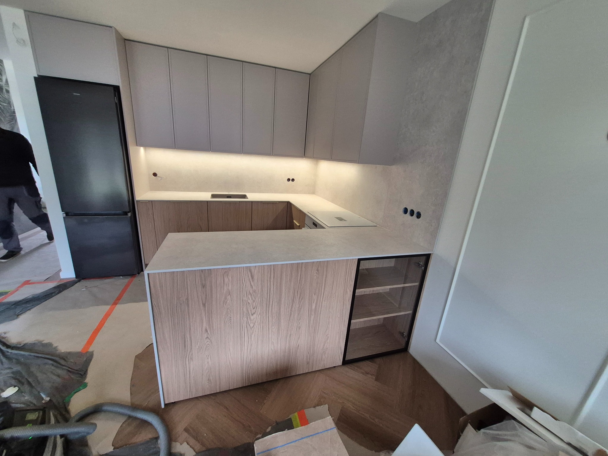 Nowoczesna kuchnia w trakcie montażu: szare szafki górne, drewnopodobny blat i fronty dolne, widoczne zabezpieczenia podłogi i sprzęt budowlany, fragment sylwetki osoby w tle.