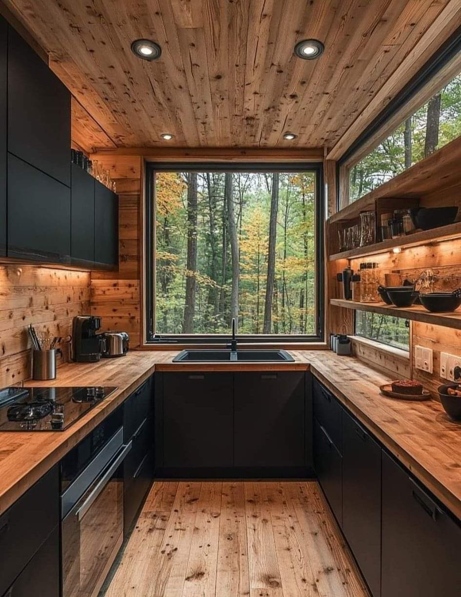 Wnętrze kuchni w stylu rustykalnym z drewnianym wykończeniem ścian, sufitu i podłogi, czarne szafki i blat z jasnego drewna, duże okno z widokiem na las.