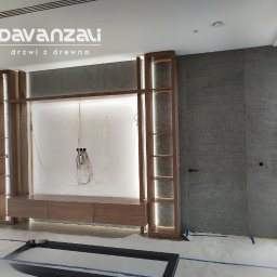 Davanzali -drzwi z drewna - Drewniana zabudowa ściany z ukrytymi drzwiami i podświetlanymi półkami. Widoczne kable i zabezpieczenia podłogi. Minimalistyczny design w szarościach i brązach.