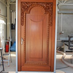 Davanzali -drzwi z drewna - Ręcznie rzeźbione, drewniane drzwi w odcieniach brązu, zamontowane w metalowej ramie z drewnianymi rozpórkami, w warsztacie stolarskim. Widoczny detal rzeźbienia.