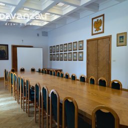 Davanzali -drzwi z drewna - Sala konferencyjna z długim drewnianym stołem i krzesłami. Na ścianie drewniane drzwi z dekoracyjnym wzorem i godło. Parkiet w jodełkę.
