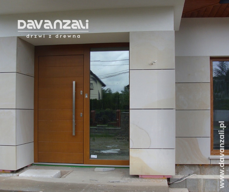 Nowoczesne drzwi wejściowe z drewna z długim, pionowym uchwytem, osadzone w jasnej, kamiennej elewacji z dużym przeszkleniem obok. Widoczny fragment domu w odbiciu.