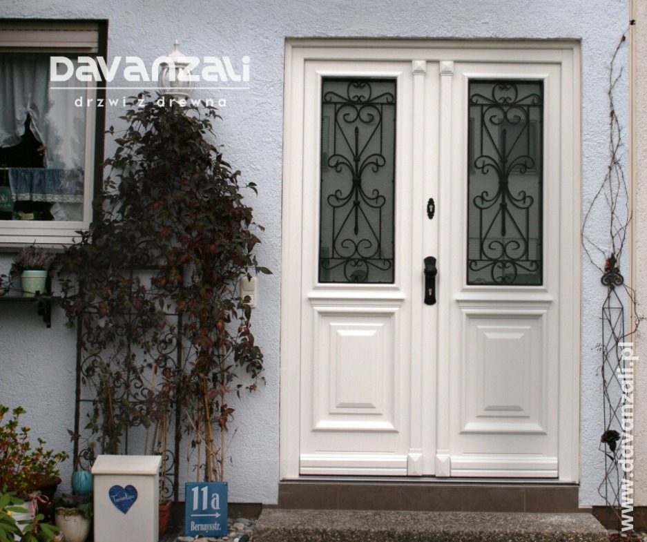 Białe, dwuskrzydłowe drzwi wejściowe z ozdobnymi, czarnymi kratami w szybach, roślina pnąca po lewej stronie, skrzynka na listy i tabliczka z numerem domu.