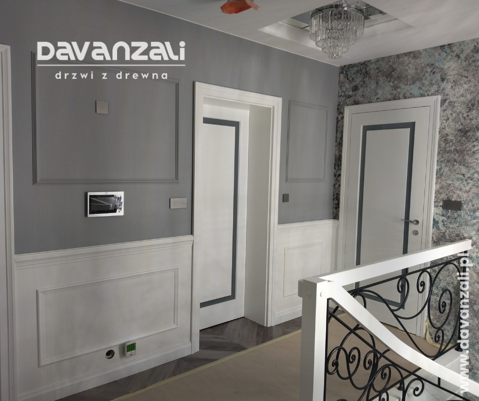 Nowoczesne, białe drzwi wewnętrzne z szarą obwódką w eleganckim holu. Widoczne dekoracyjne panele ścienne i kryształowy żyrandol. Ujęcie z perspektywy balustrady.
