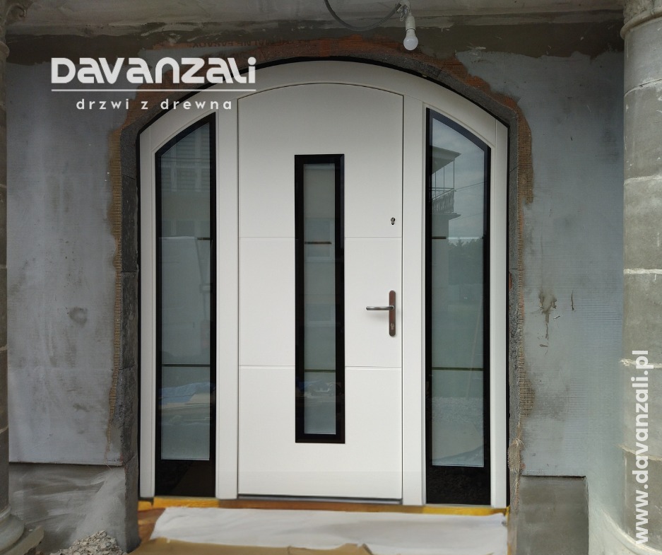 Białe drzwi wejściowe z przeszkleniami bocznymi, czarne ramki, łukowe nadproże, surowy mur, widoczny montaż, logo producenta u góry.