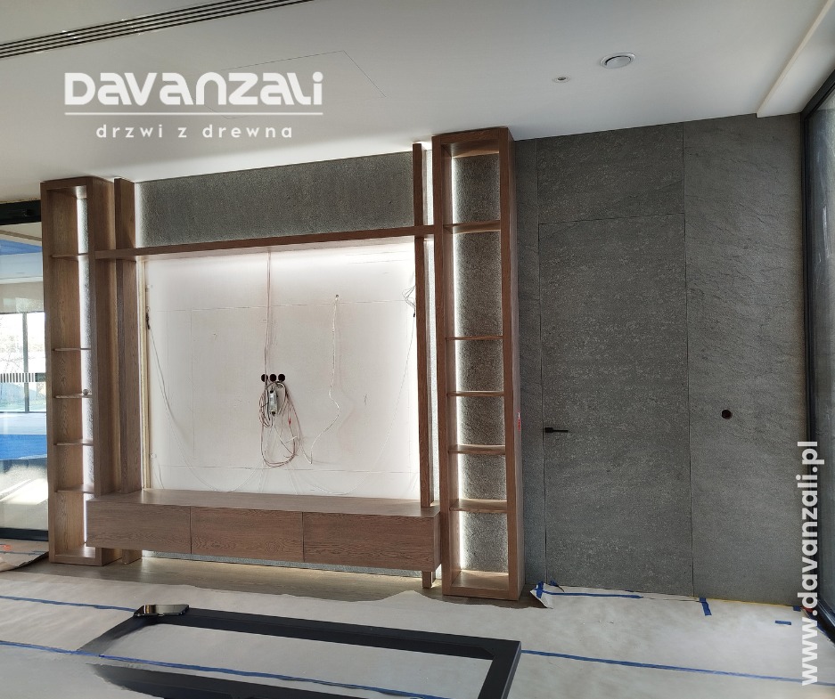 Drewniana zabudowa ściany z ukrytymi drzwiami i podświetlanymi półkami. Widoczne kable i zabezpieczenia podłogi. Minimalistyczny design w szarościach i brązach.