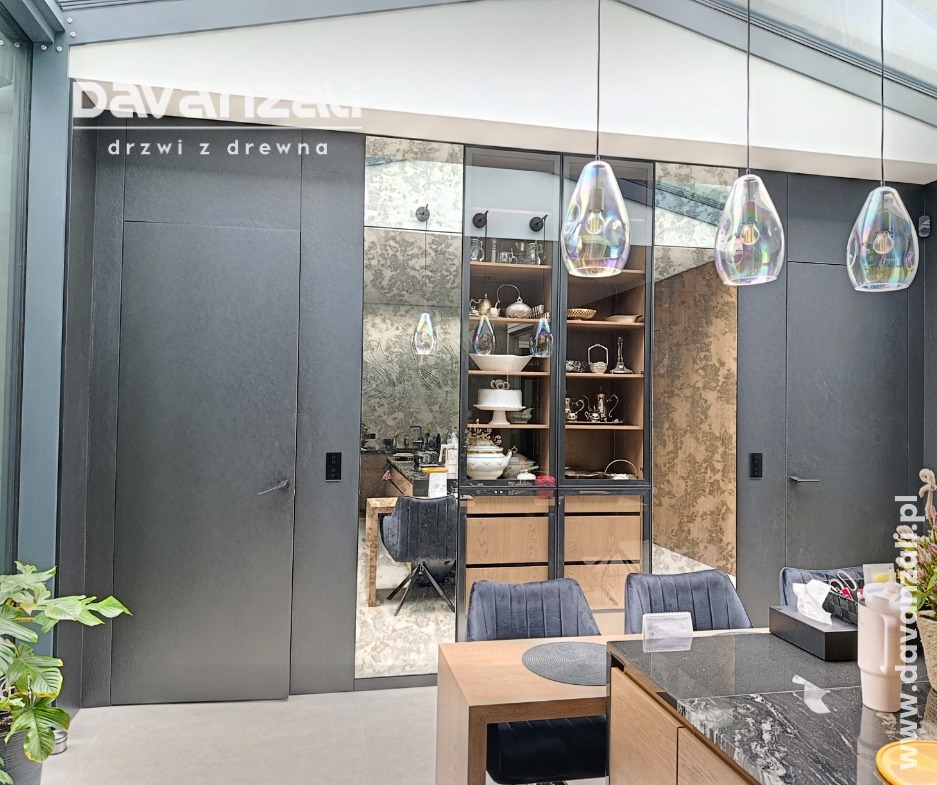 Nowoczesne, szare drzwi wewnętrzne z ukrytą ościeżnicą, wkomponowane w ścianę z lustrami i witryną. Nad stołem wiszą trzy szklane lampy.