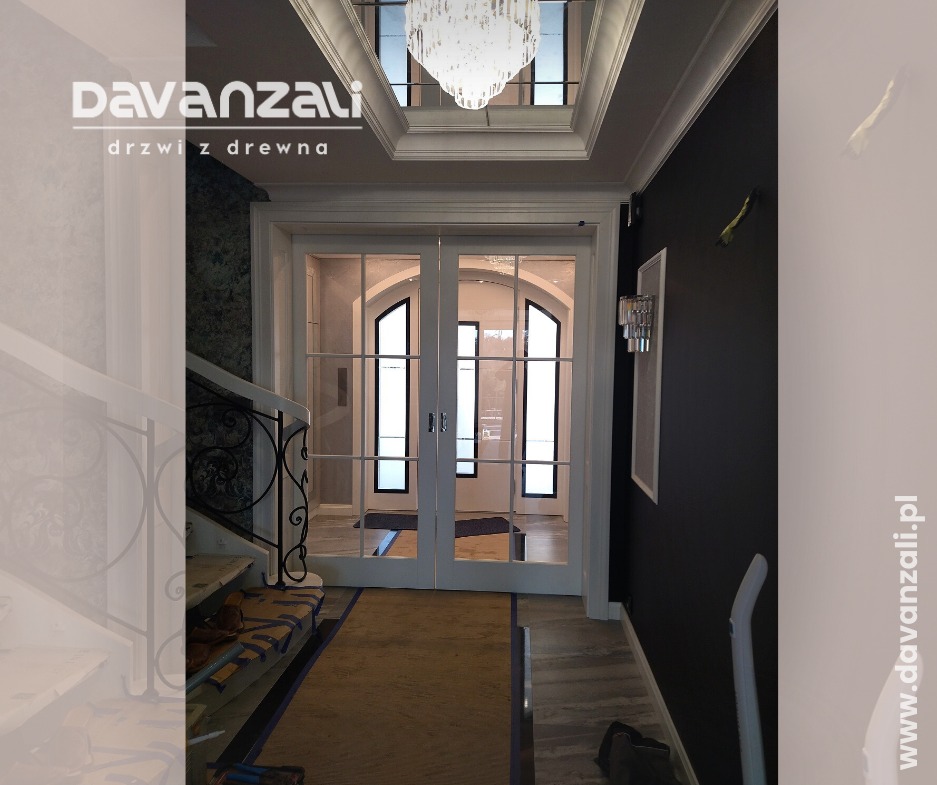 Eleganckie, białe drzwi wewnętrzne z przeszkleniami, prowadzące do jasnego pomieszczenia. Widoczne schody z ozdobną balustradą i kryształowy żyrandol.