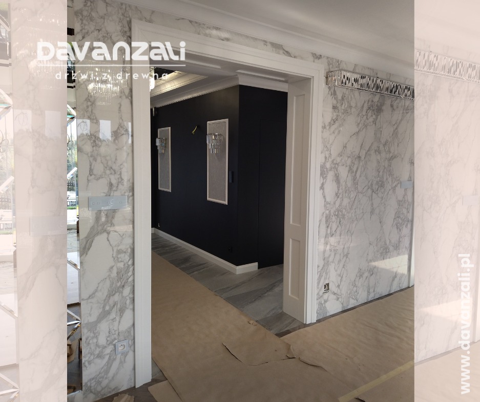 Białe, drewniane drzwi wewnętrzne z ościeżnicą, wkomponowane w marmurowe wykończenie ściany. Widok na ciemny korytarz z oświetleniem i marmurową podłogą.