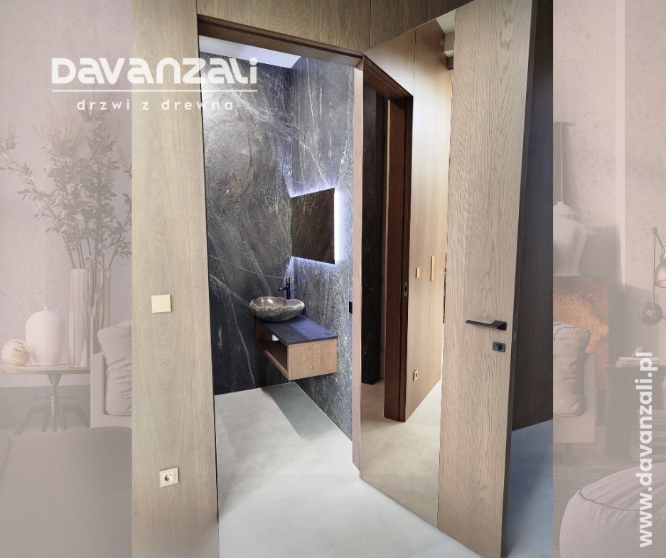 Nowoczesne, drewniane drzwi wewnętrzne z czarną klamką, otwarte, ukazujące łazienkę z kamienną umywalką i podświetlanym lustrem. Minimalistyczny design.