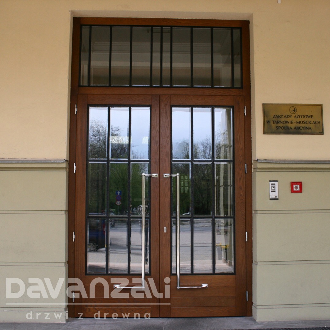 Dwuskrzydłowe drzwi wejściowe z drewna i szkła, z długimi, srebrnymi uchwytami, zamontowane w budynku z tablicą informacyjną po prawej stronie.