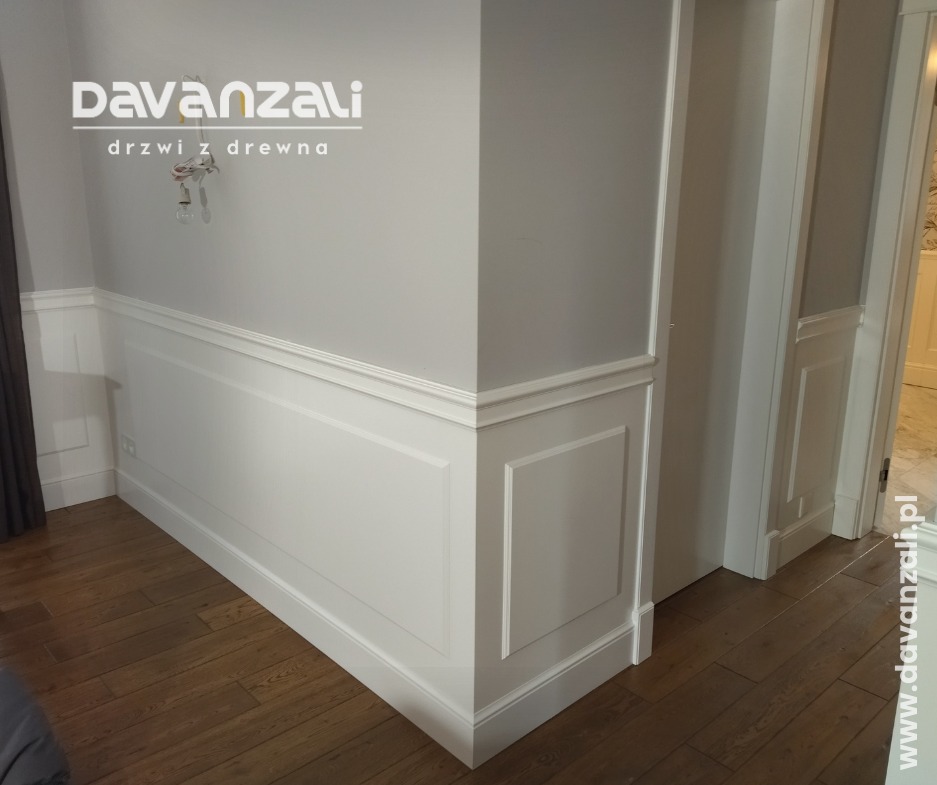 Białe, drewniane panele ścienne z listwami przypodłogowymi i fragmentem drzwi wewnętrznych w jasnym wnętrzu. Widoczna podłoga z drewnianych desek.