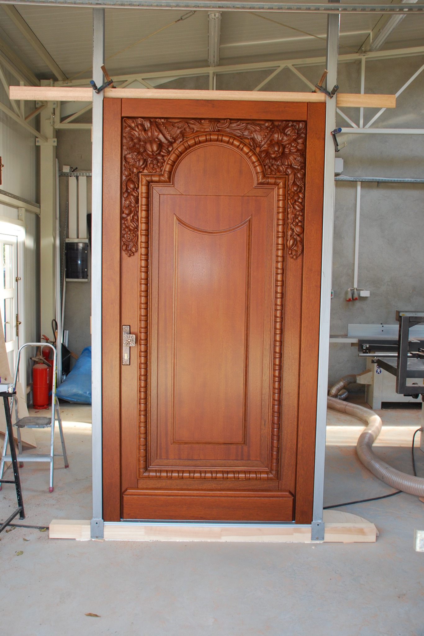 Ręcznie rzeźbione, drewniane drzwi w odcieniach brązu, zamontowane w metalowej ramie z drewnianymi rozpórkami, w warsztacie stolarskim. Widoczny detal rzeźbienia.