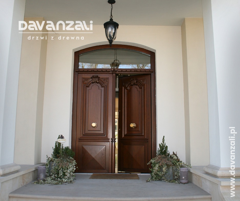 Ciemne, drewniane drzwi wejściowe do budynku z kolumnami, częściowo otwarte, z dekoracyjnymi roślinami po bokach. Nad drzwiami łukowe okno i wisząca lampa.