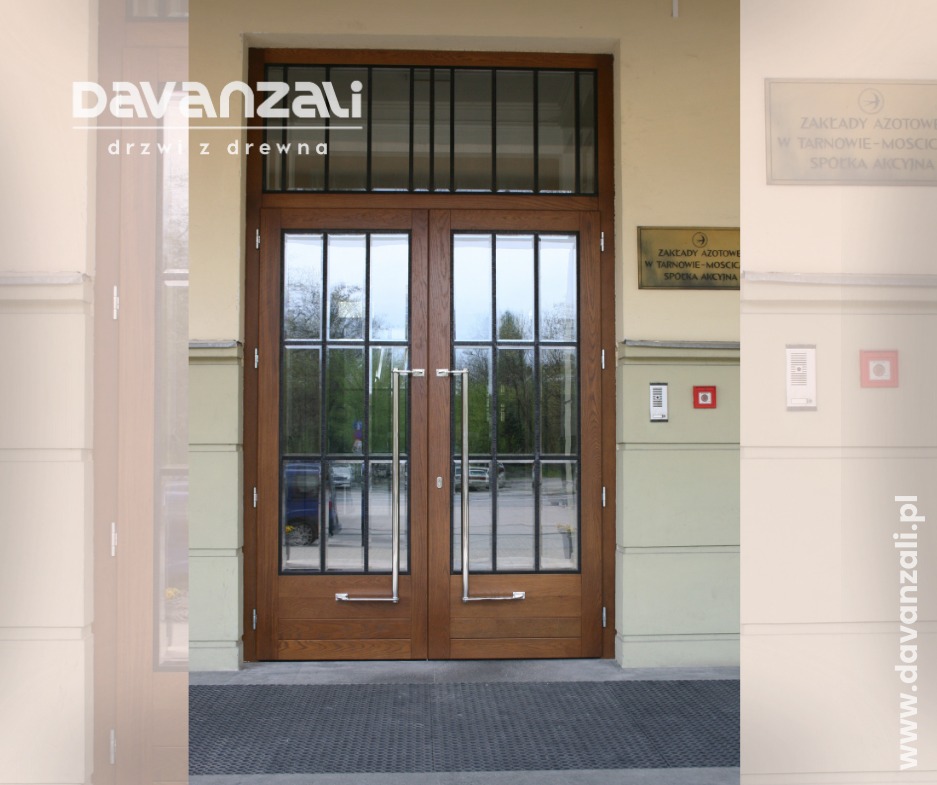 Dwuskrzydłowe, drewniane drzwi wejściowe z przeszkleniami i metalowymi szprosami, z długimi, pionowymi uchwytami, zamontowane w budynku z beżową elewacją.