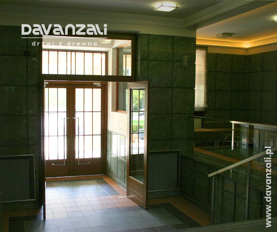 Drewniane, dwuskrzydłowe drzwi z przeszkleniami w holu wykończonym płytkami w odcieniach zieleni i brązu. Otwarta pojedyncza furtka po prawej stronie.