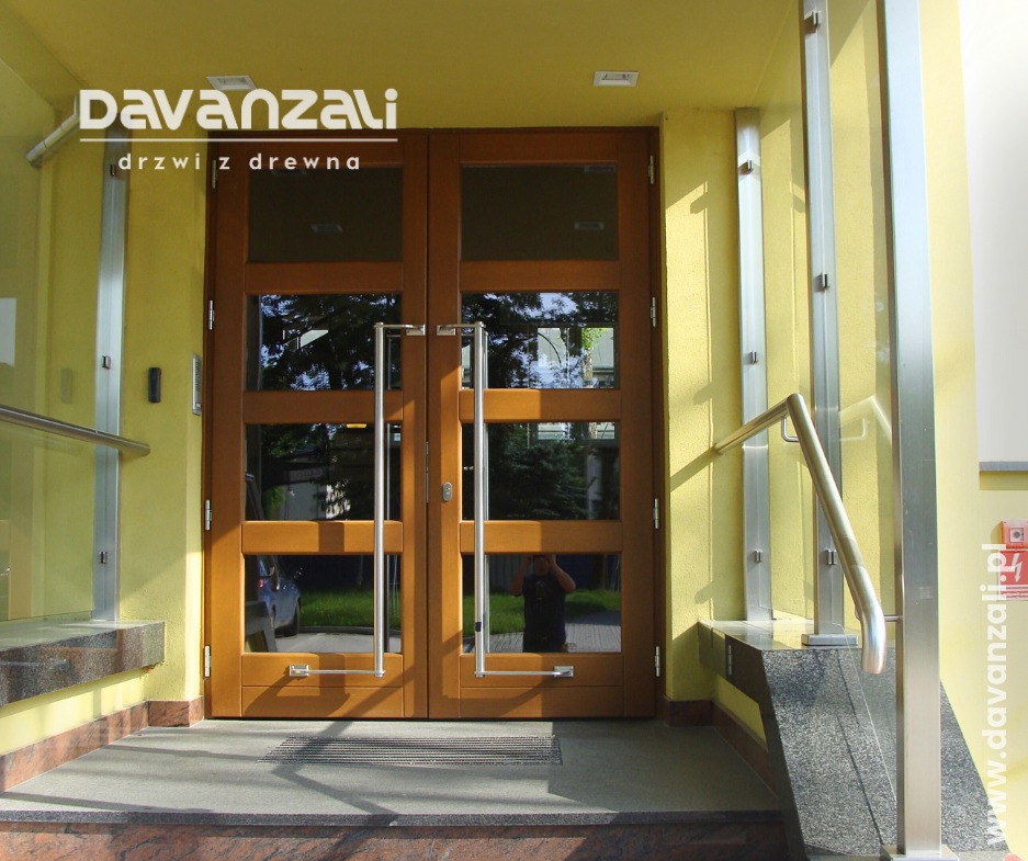 Dwuskrzydłowe drzwi wejściowe z drewna z przeszkleniami, stalowe uchwyty, widoczne odbicie otoczenia w szybach, żółta elewacja budynku.