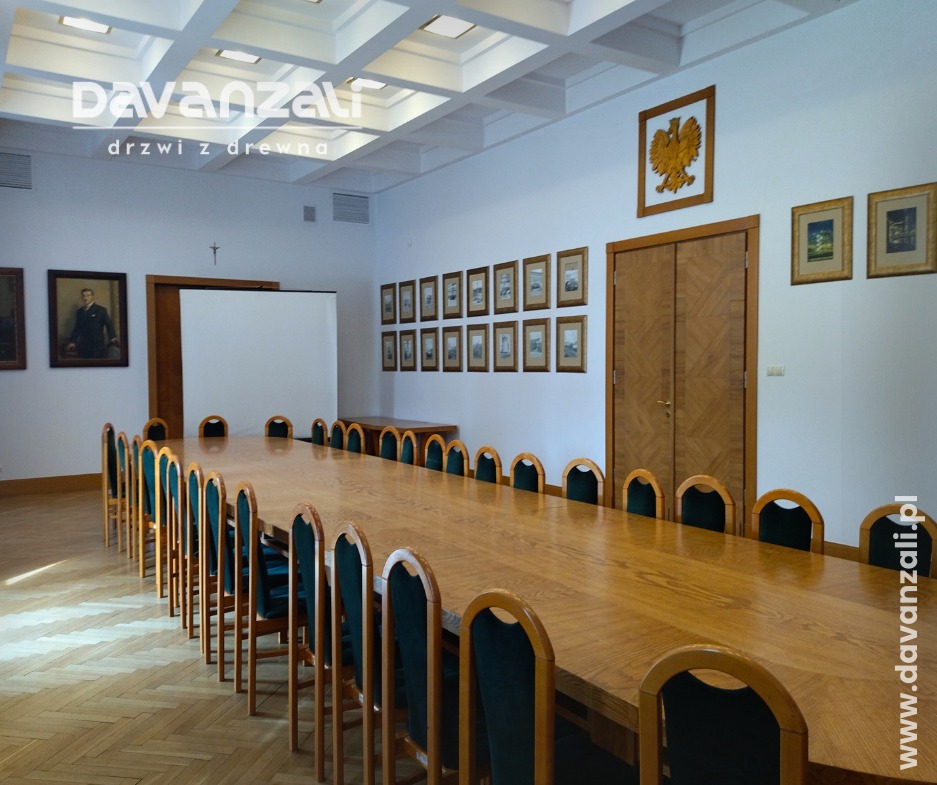Sala konferencyjna z długim drewnianym stołem i krzesłami. Na ścianie drewniane drzwi z dekoracyjnym wzorem i godło. Parkiet w jodełkę.