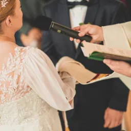 Panna Młoda w sukni ślubnej z koronkowymi plecami podczas ceremonii ślubnej, mikrofon i ksiądz w kadrze. Ujęcie zza ramienia, skupione na detalu sukni.