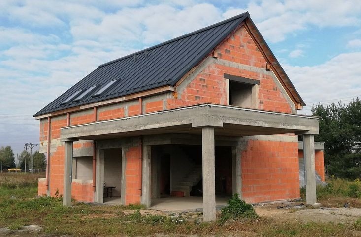 Niedokończony budynek jednorodzinny z czerwonej cegły z szarym dachem i trzema oknami dachowymi, widok z zewnątrz na parter z betonowymi filarami i zadaszeniem, w tle drzewa i niebo.