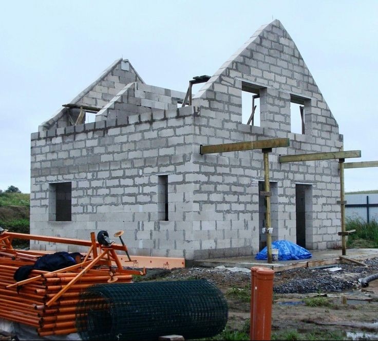 Surowy stan domu z szarego betonu komórkowego z widocznymi otworami okiennymi i drzwiowymi, wsparty drewnianymi stemplami, z pomarańczowymi rusztowaniami i zwojem siatki ogrodzeniowej w tle.