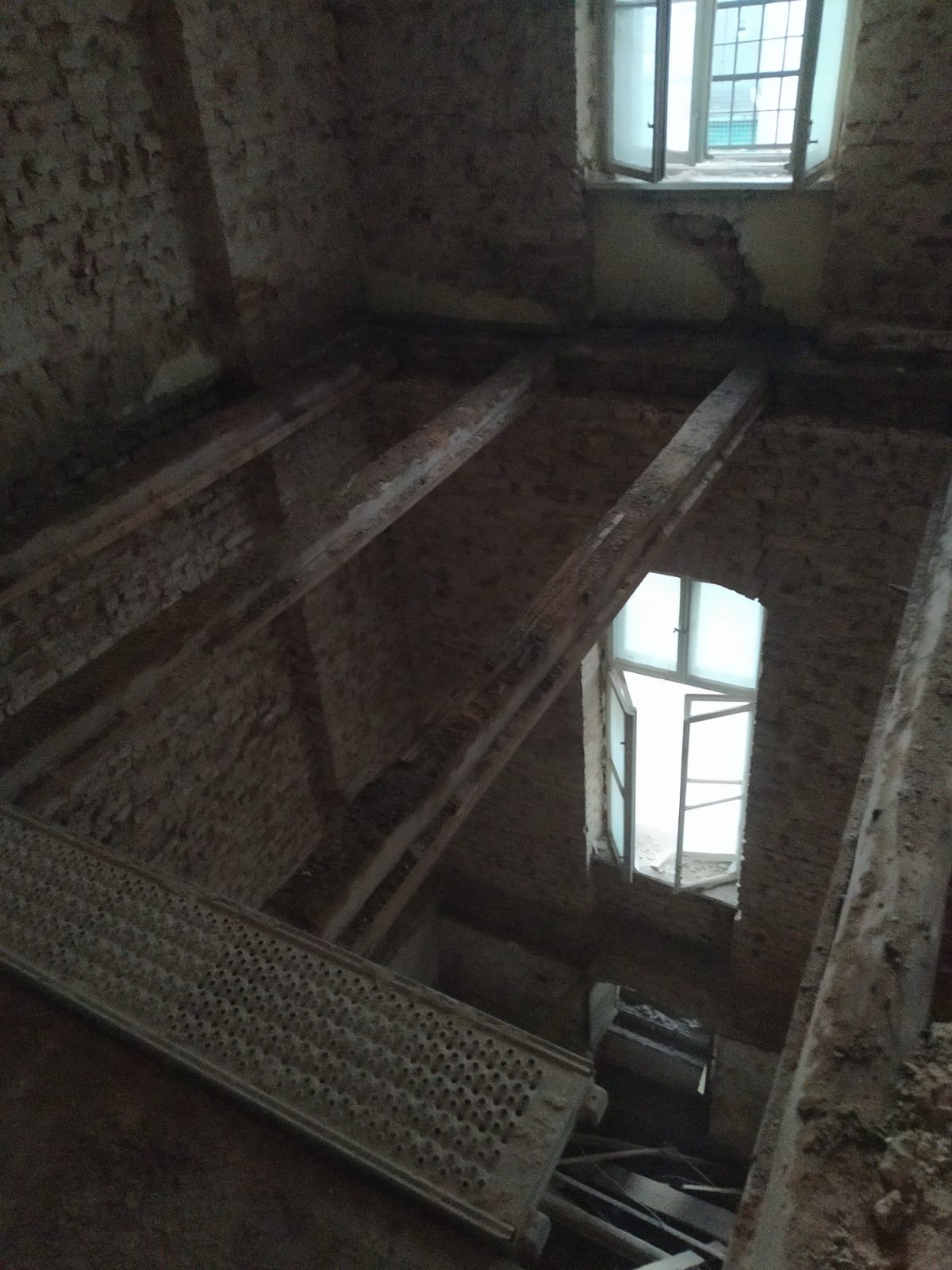 Wnętrze budynku w trakcie rozbiórki, widoczne ceglane ściany, drewniane belki stropowe, okna z otwartymi skrzydłami i metalowa platforma. Fragmenty gruzu i odpadów budowlanych na podłodze.