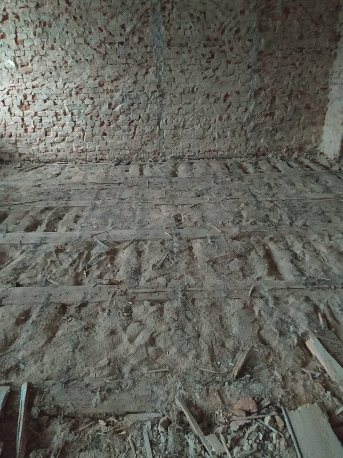 Wnętrze zniszczonego budynku z odsłoniętą ceglaną ścianą i gruzem na podłodze, pozostałości po demontażu podłogi.