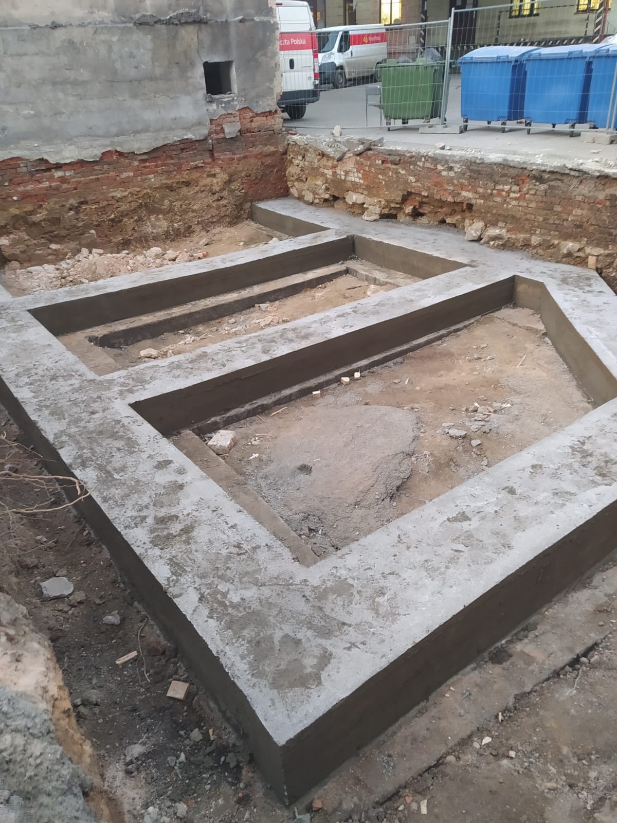 Wylany fundament pod budynek z widocznymi fragmentami starej ceglanej ściany, na zewnątrz budowy zaparkowane busy poczty polskiej.