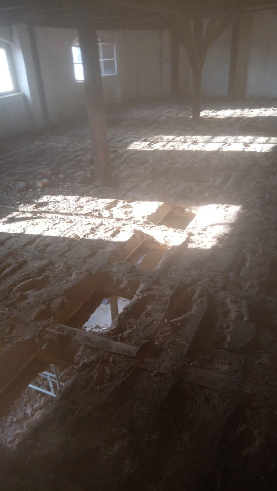 Wnętrze budynku w trakcie rozbiórki, widoczne drewniane słupy, okna i gruz na podłodze z odsłoniętymi fragmentami konstrukcji.