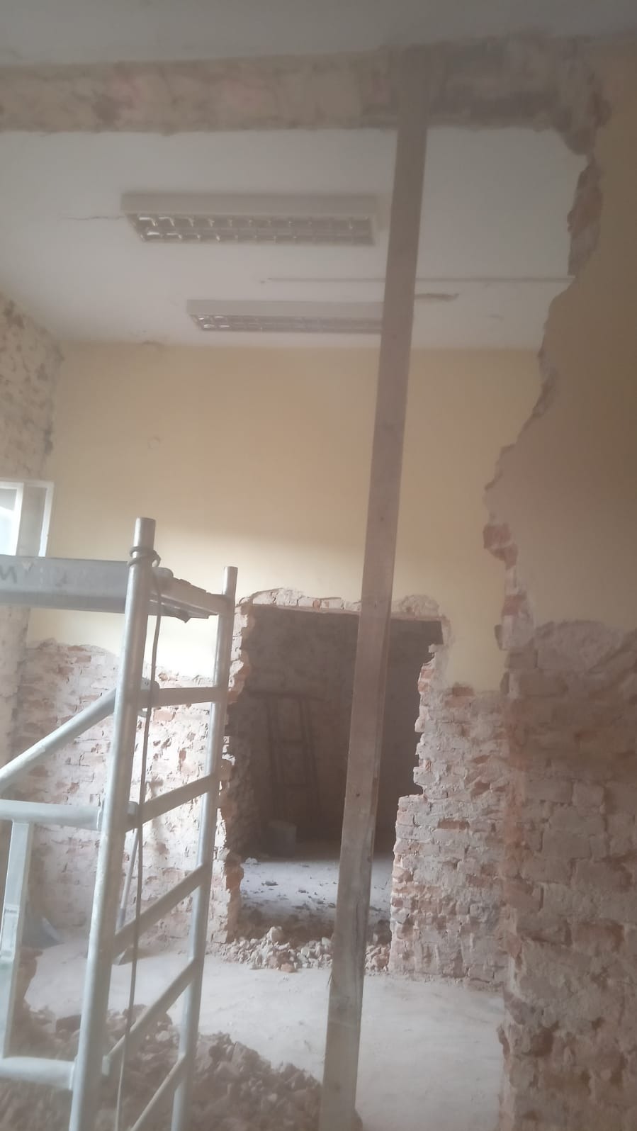 Wnętrze w trakcie prac remontowych: częściowo usunięte ściany z cegły, widoczne rusztowanie, gruz na podłodze, odsłonięte oświetlenie sufitowe.