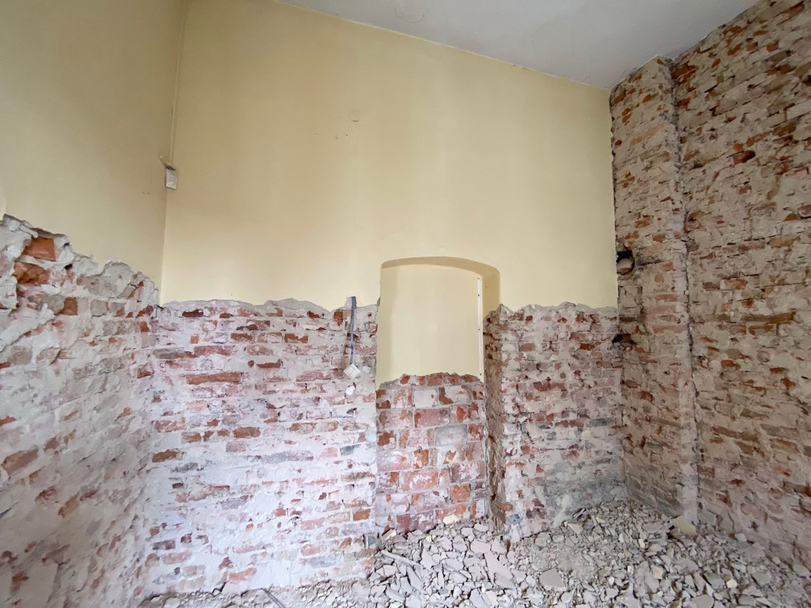 Wnętrze pomieszczenia w trakcie prac remontowych, z częściowo usuniętym tynkiem odsłaniającym ceglaną ścianę, widoczne pozostałości gruzu na podłodze, fragment otworu drzwiowego w ścianie.