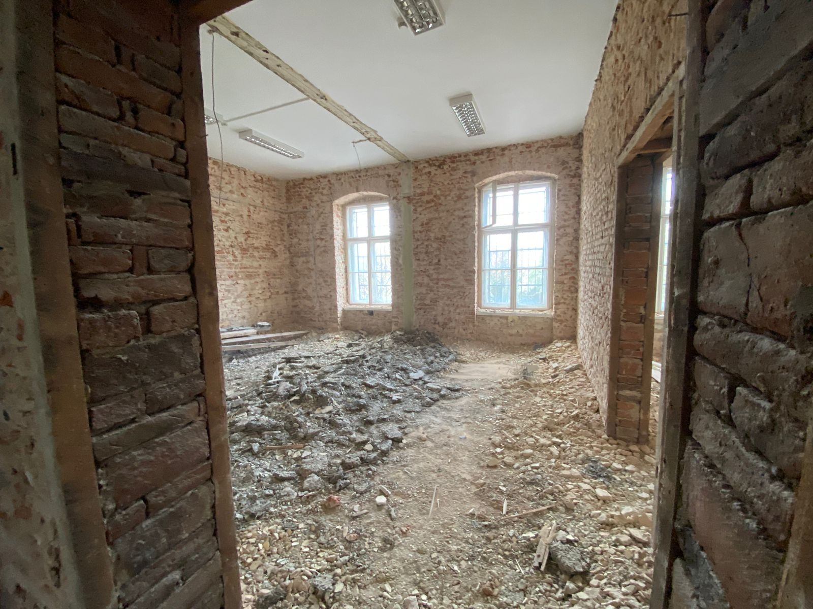 Wnętrze pomieszczenia z odsłoniętą cegłą na ścianach, gruzem na podłodze i dwoma oknami, widok zza framugi drzwi.