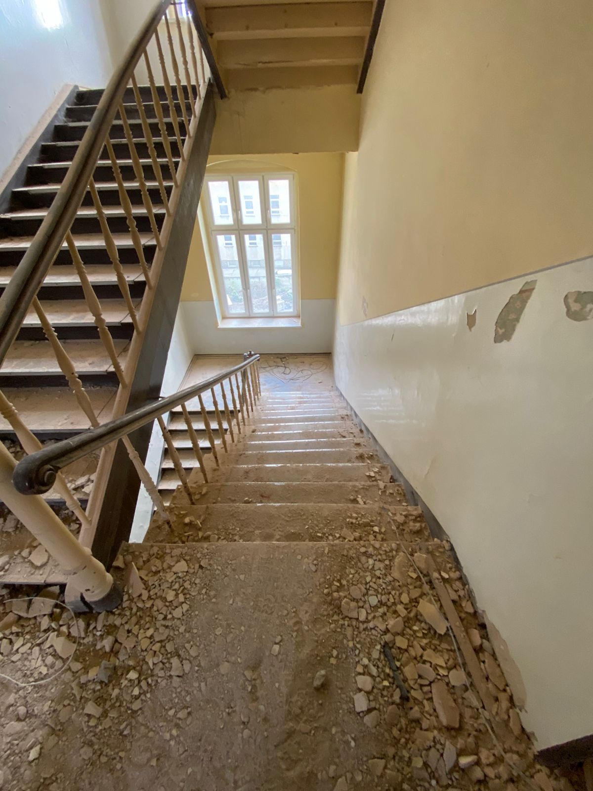 Klatka schodowa w trakcie remontu: gruz na schodach, uszkodzone ściany, widoczne stare drewniane balustrady, okno w tle.