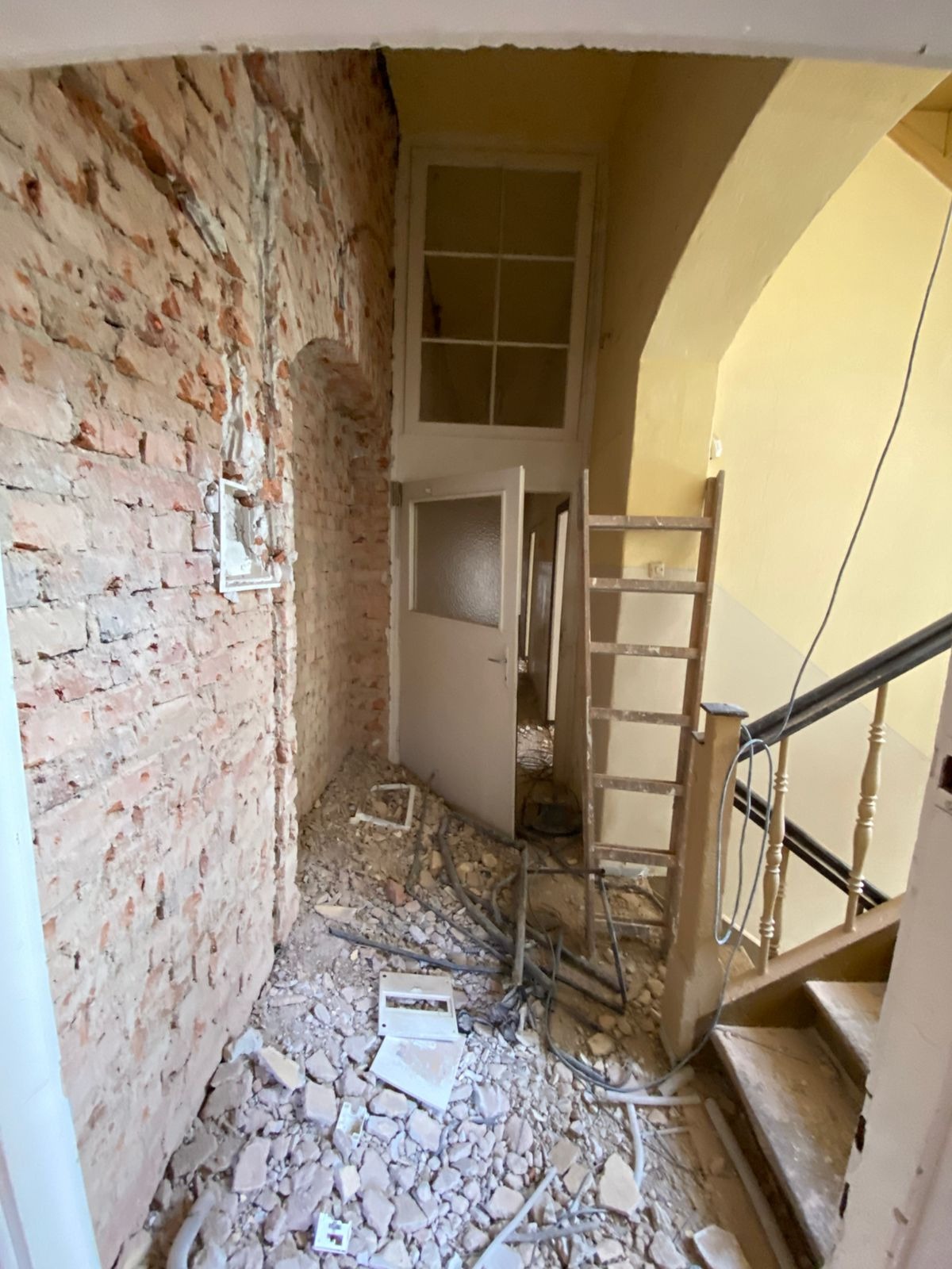 Wnętrze budynku w trakcie prac: odsłonięta ceglana ściana, gruzowisko na podłodze z widocznymi przewodami, otwarta biała skrzynka elektryczna, drewniana drabina oparta o ścianę przy schodach.