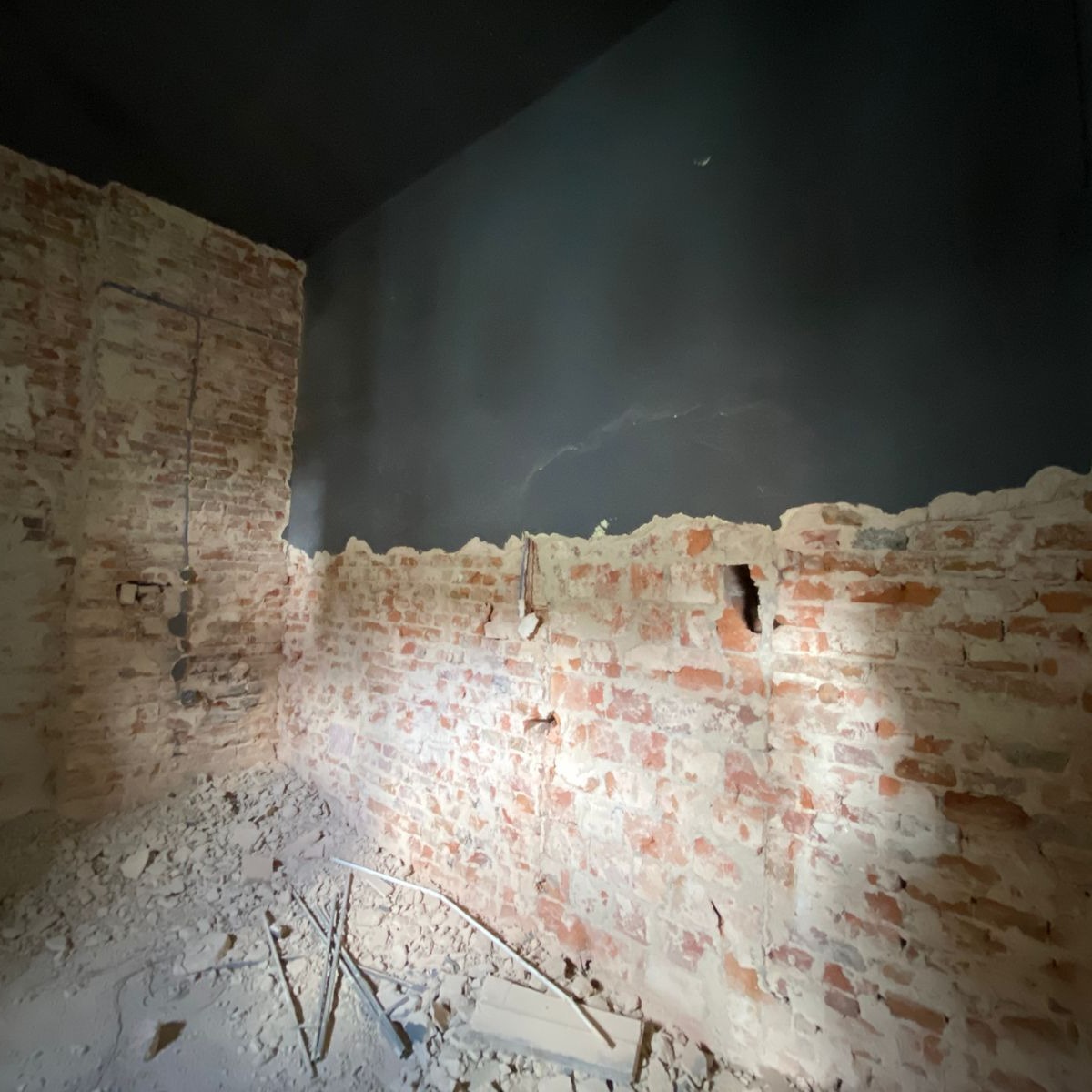 Wnętrze pomieszczenia w trakcie prac rozbiórkowych: częściowo otynkowana ceglana ściana z widocznymi przewodami, gruz na podłodze i ciemny sufit.