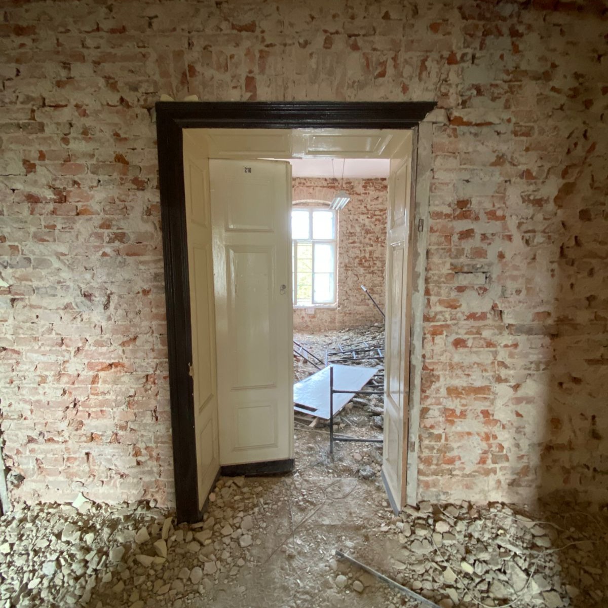 Ceglana ściana z otwartymi drzwiami do zdemolowanego pokoju, gruz na podłodze, widoczne pozostałości po pracach rozbiórkowych.
