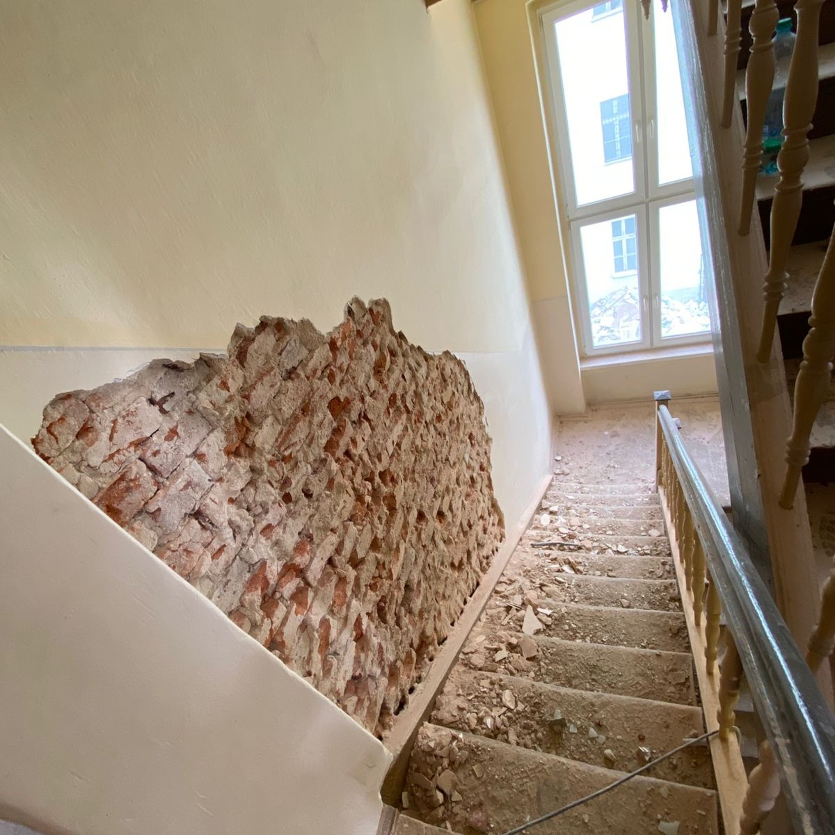 Fragmentalnie wyburzona ściana z czerwonej cegły na klatce schodowej, widok z góry na schody pokryte gruzem, okno w tle.