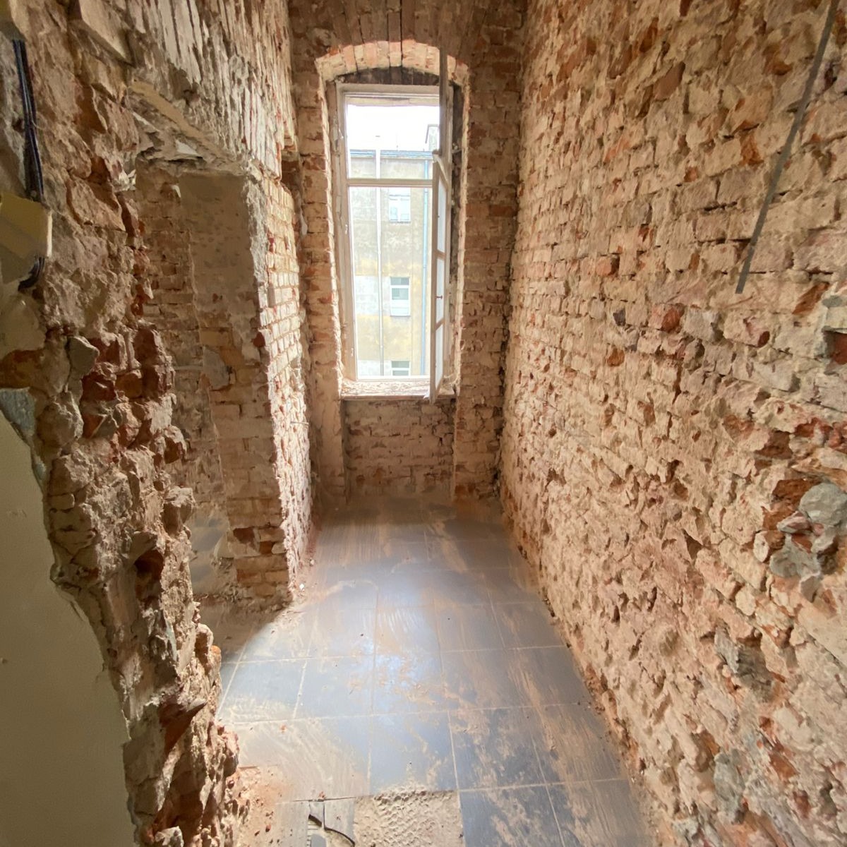 Wnętrze pomieszczenia w trakcie remontu: odsłonięte ceglane ściany z ubytkami tynku, fragmenty instalacji elektrycznej, okno z widokiem na budynek, szare płytki podłogowe pokryte pyłem.