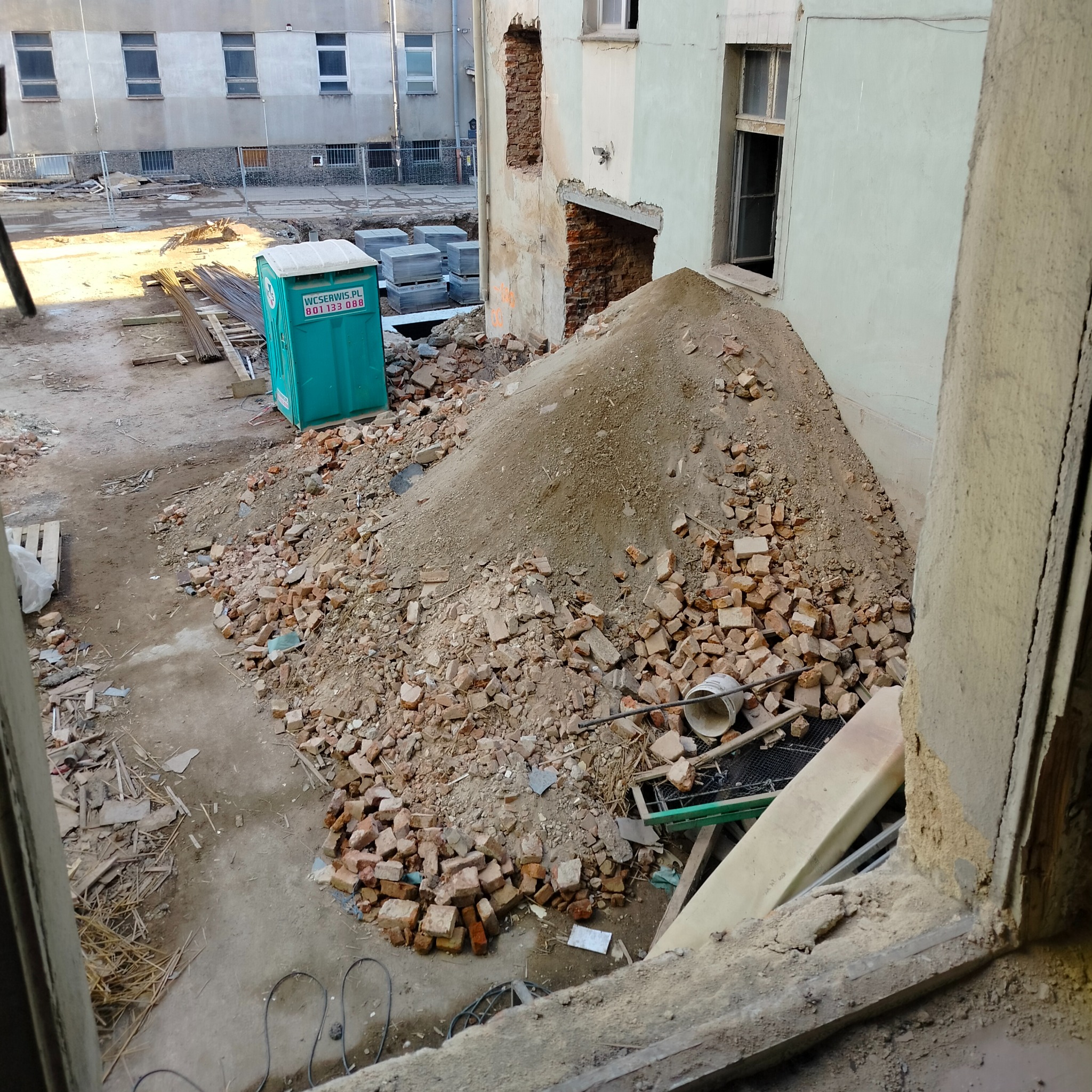 Widok z okna na plac budowy po wyburzeniach, sterta gruzu ceglanego, przenośna toaleta TOI TOI w tle, elementy konstrukcji budynku z odsłoniętą cegłą.